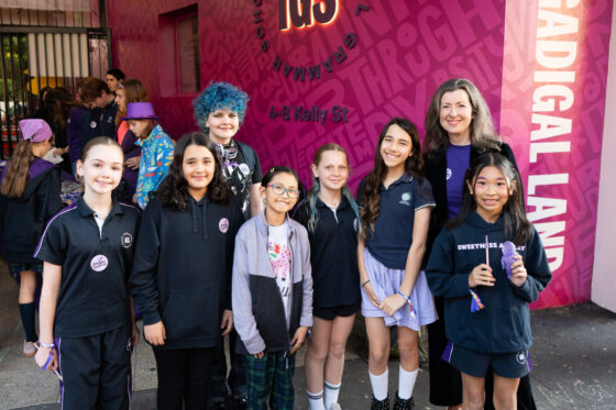 A group of children and an adult stand smiling together in front of a bright pink wall with writing, proudly wearing Colours of Courage badges and casual clothes.