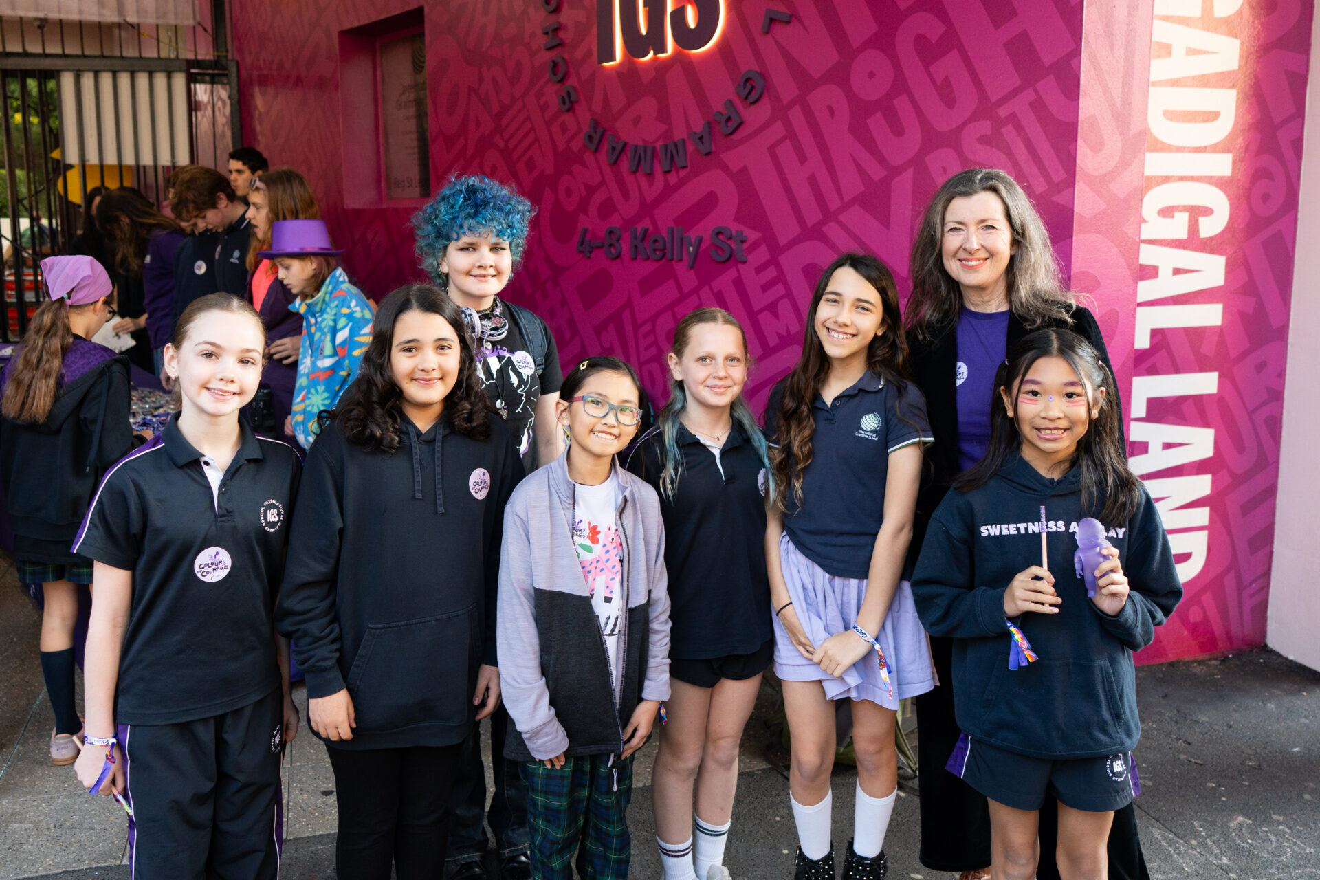 A group of children and an adult stand smiling together in front of a bright pink wall with writing, proudly wearing Colours of Courage badges and casual clothes.