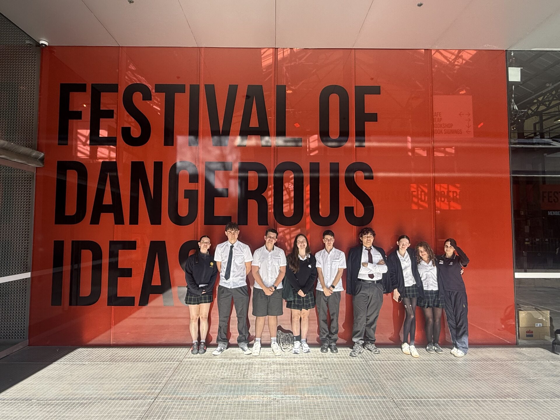 A group of ten students in school uniforms stands in front of a large red wall with the words Festival of Dangerous Ideas in bold black letters.