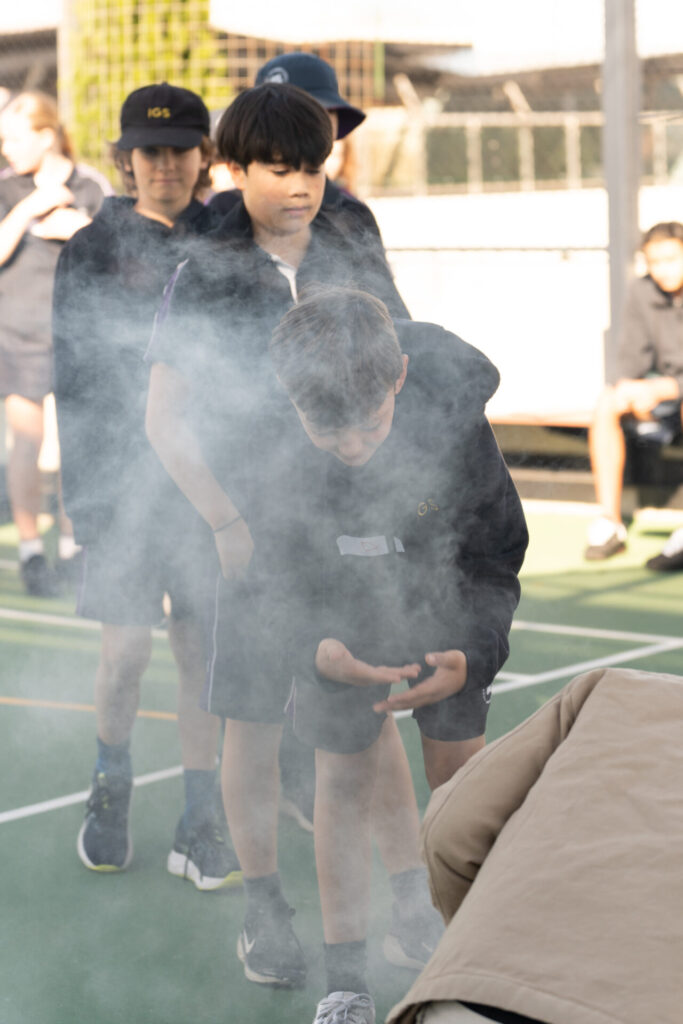 Several children in sports kits walk in a line through a cloud of smoke on an outdoor court, their authentic voices echoing with excitement. One boy in front bends slightly forward, holding something in his hands, as if learning from the experience. Other children and adults are visible in the background.