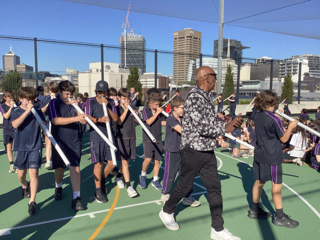 A group of school children, each bringing their authentic voices, play white tube instruments outdoors on a sports court, led by an adult in sunglasses. City buildings and cranes are visible in the background, creating a vibrant setting for hands-on learning.