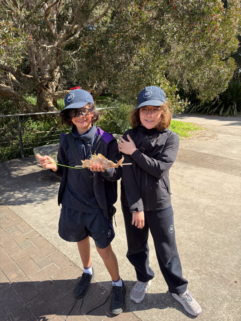 Two children wearing navy hats and black outfits stand outdoors on a sunny day, immersed in learning through play. One child holds a large stick with leaves, while the other smiles and rests a hand on their friend’s shoulder. Trees and pavement are in the background, capturing an authentic moment of friendship and authentic voices.
