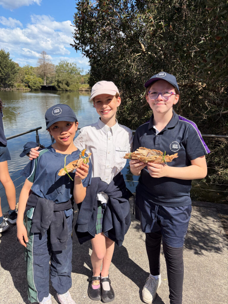 Three children stand together by a pond, smiling at the camera. Two are holding leaves and sticks as part of their learning adventure. All are wearing casual clothing and hats. The background shows water, trees, and a partly cloudy sky.