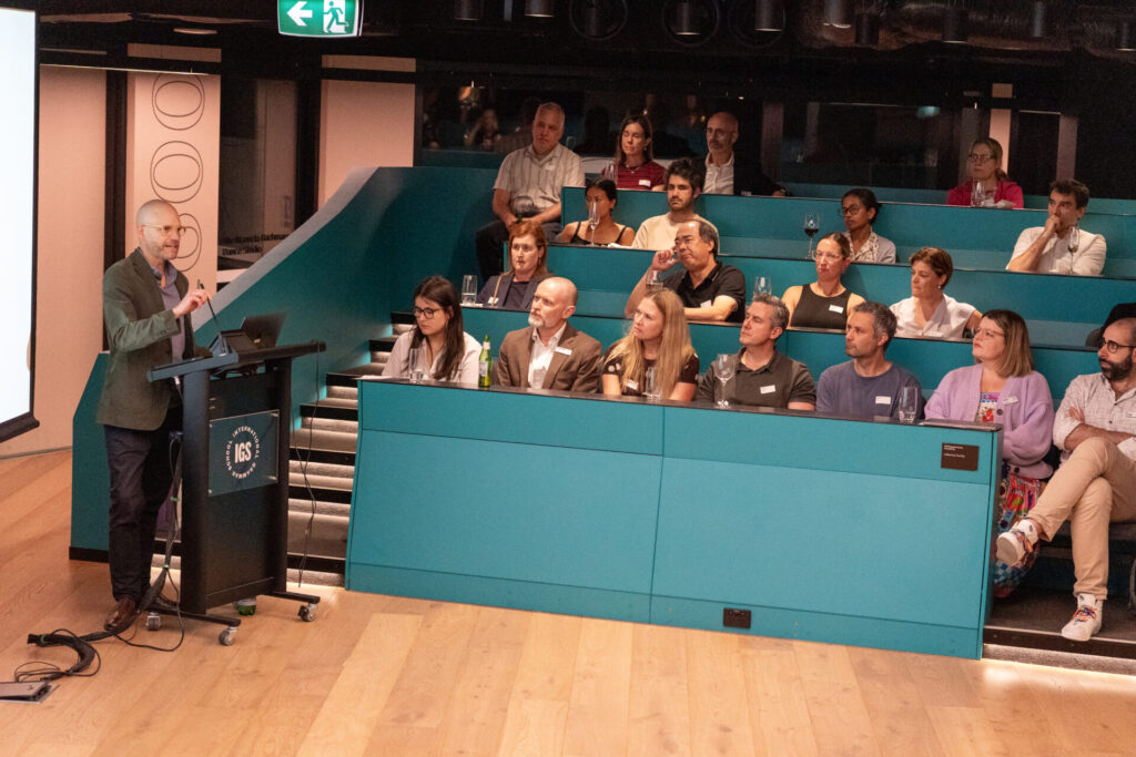 A speaker presents at a lectern to a seated audience in a modern lecture theatre with teal seating and wooden flooring. The audience listens attentively, some taking notes or looking at the screen, as the presentation explores topics such as behaviour and emotional regulation.