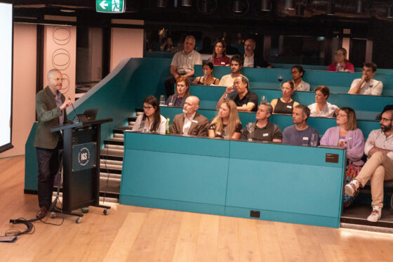 A speaker presents at a lectern to a seated audience in a modern lecture theatre with teal seating and wooden flooring. The audience listens attentively, some taking notes or looking at the screen, as the presentation explores topics such as behaviour and emotional regulation.