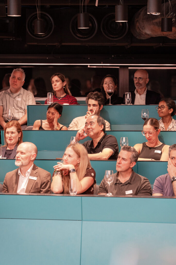 A diverse group of people sits in tiered rows of turquoise seats, attentively listening to an event or presentation that explores topics such as behaviour and emotional regulation, with glasses and notebooks in front of some attendees.