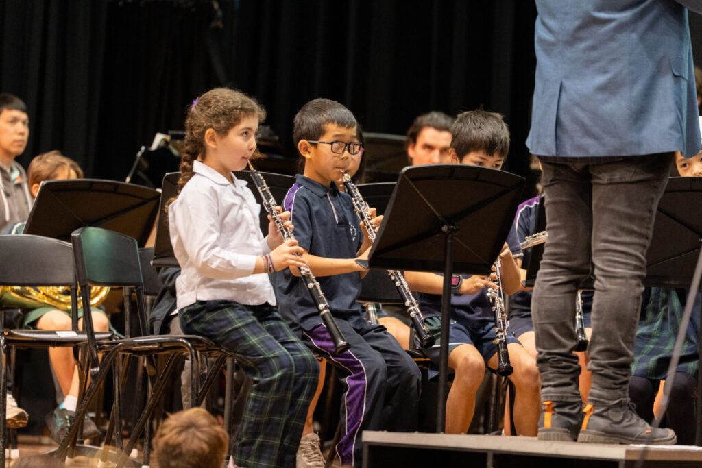 Children in school uniforms sit in chairs playing wind instruments during a band performance, highlighting the connection between music and learning. A conductor stands in front, and other musicians are visible in the background.