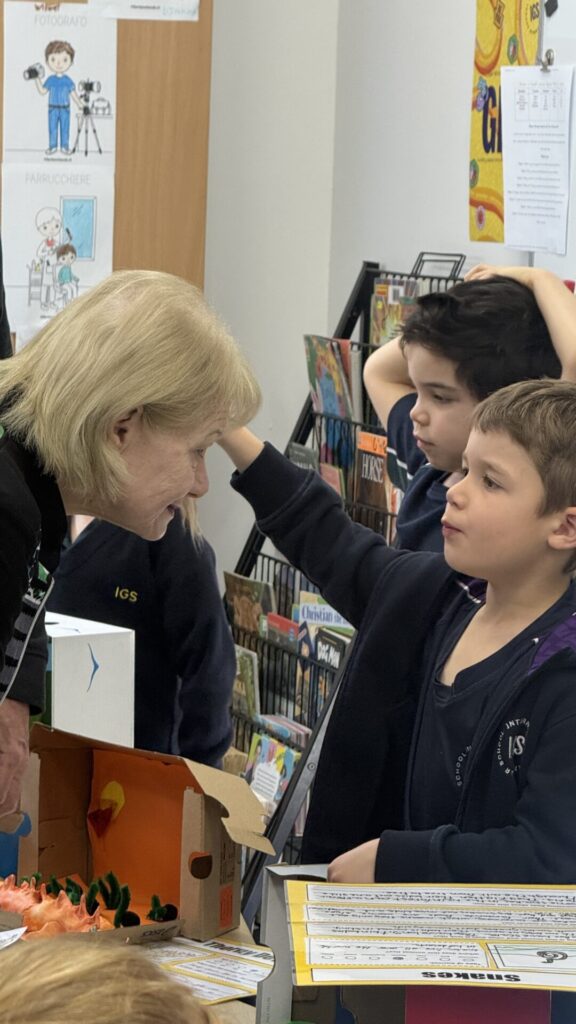A woman leans in to listen to a young boy speaking at a classroom table, fostering a sense of connection and learning, whilst another child stands nearby. Educational posters and bookshelves fill the background, capturing the spirit of Grand-friends' Day.