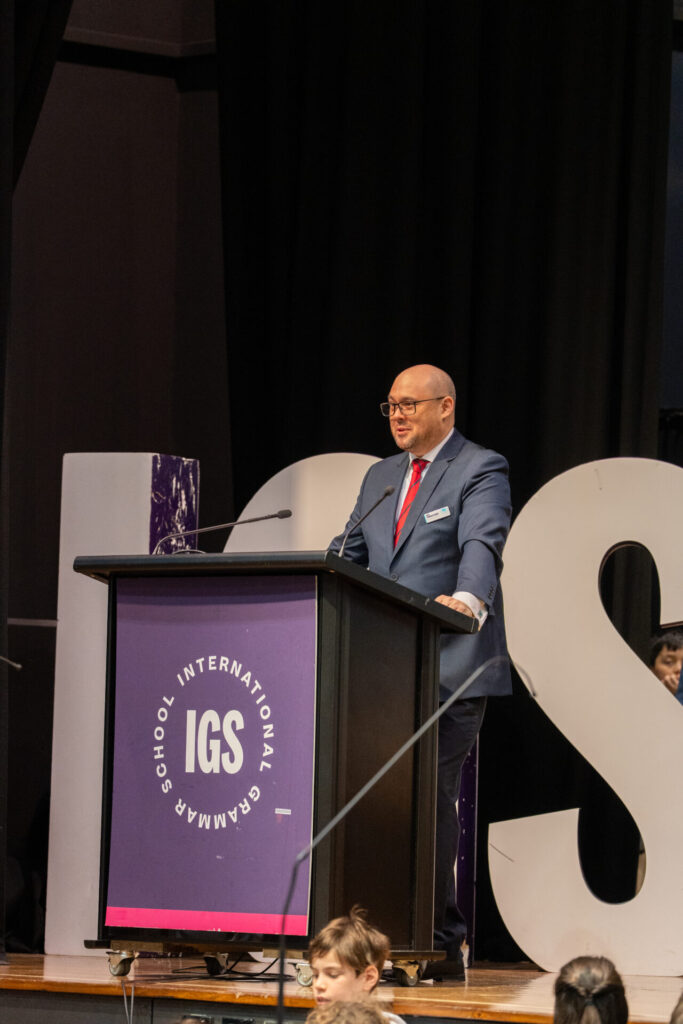 A man in a blue suit and red tie speaks at a podium labelled IGS International Grammar School, highlighting the importance of Learning and Connection. Large, white letters are in the background, and children are seated below the stage.