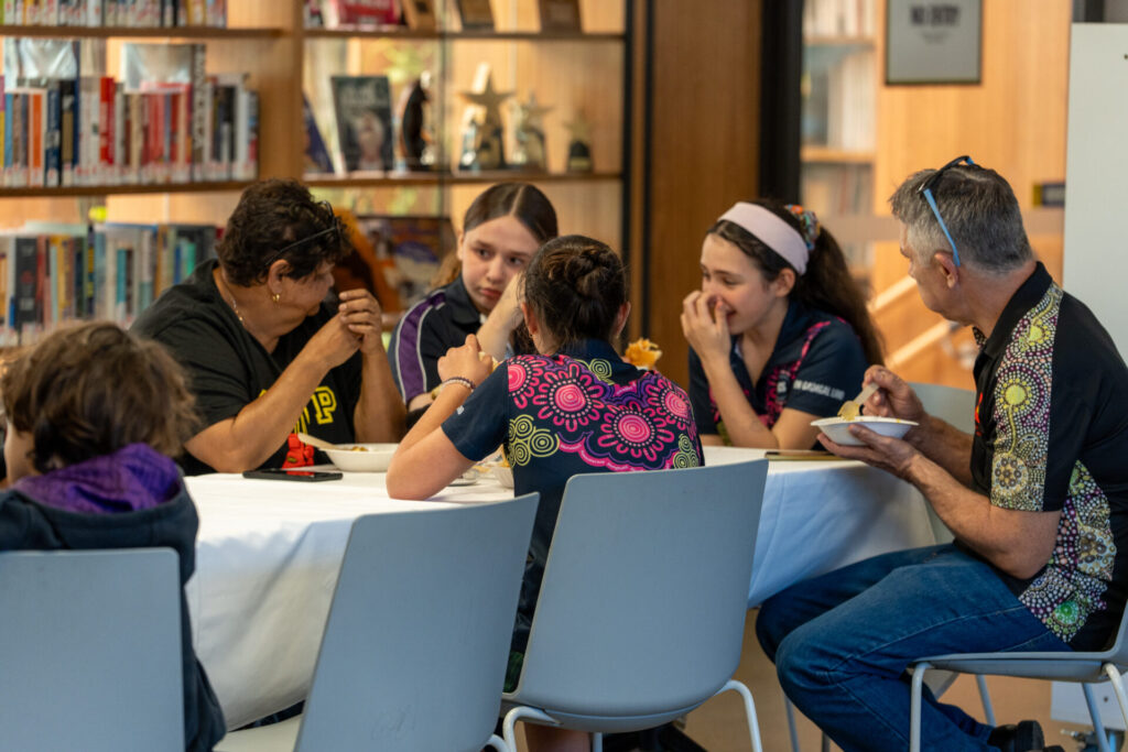 Five people sit around a table in a library, talking and eating. Three girls are engaged in conversation, while an older woman and man eat—an atmosphere that captures the spirit of Grand-friends’ Day. Surrounded by bookcases, the group enjoys meaningful connection and learning across generations.