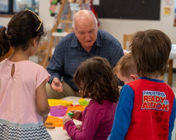 An older man interacts with four young children who are playing with toys at a table in a colourful classroom, creating a special moment of learning and connection—perfectly capturing the spirit of Grand-friends Day.