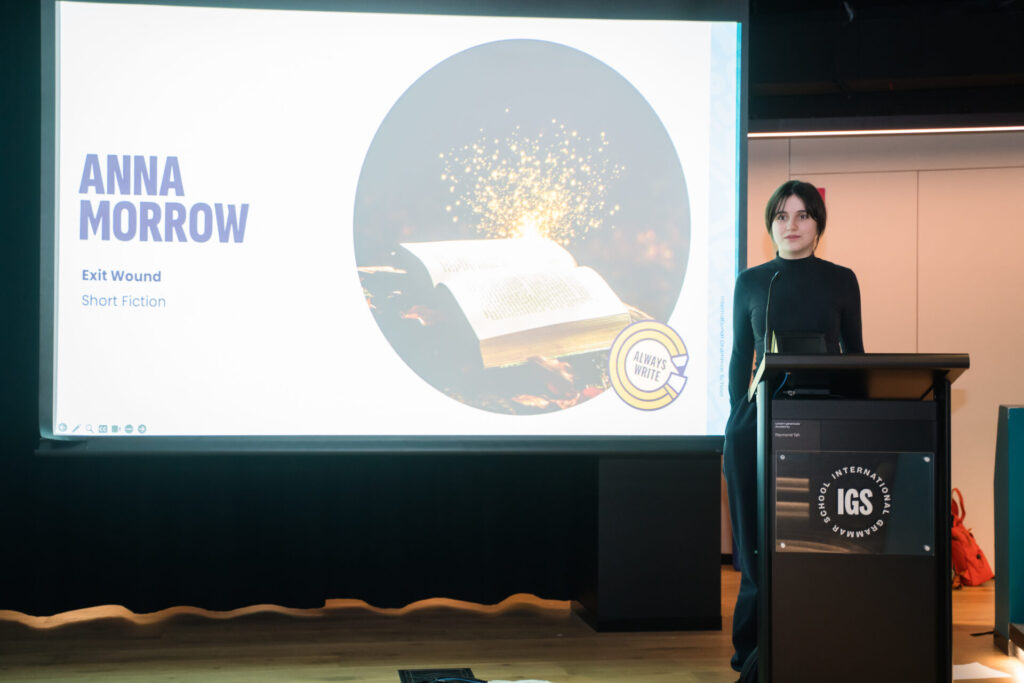 A person stands at a lectern labelled IGS beside a projected slide displaying Anna Morrow, Exit Wound, Short Fiction and an image of an open, glowing book, evoking the craft of storytelling and the intricacies of narrative arc.