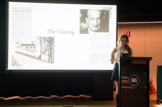 A woman gives a presentation on The Clearing, exploring elements of storytelling with a slide showing images of Martin Heidegger, his book Sein und Zeit, and a house, at a lectern labelled IGS.