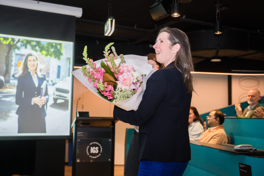 A smiling woman holds a bouquet of flowers in a modern event space. Behind her, a screen displays a photo of her, underscoring the storytelling at play as several people are seated, clapping and watching the presentation unfold.