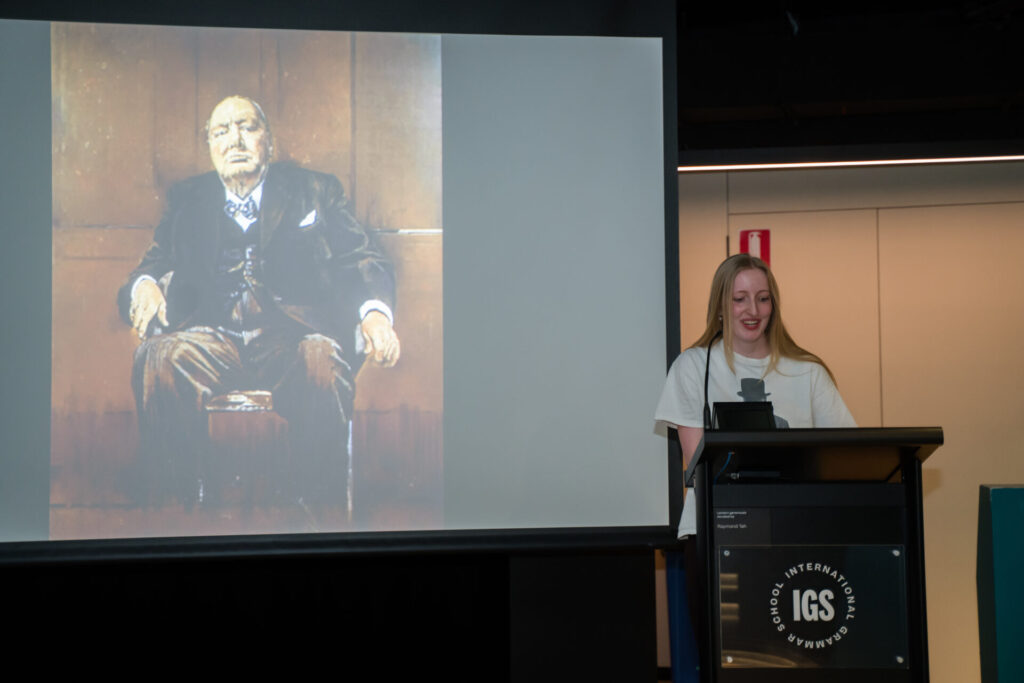 A young woman speaks at a lectern with a projected painting of an older man in a suit on a screen beside her, artfully guiding the audience through the narrative arc and illuminating the shape of his story.