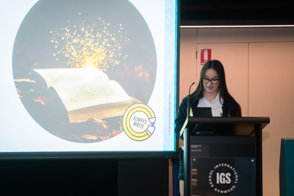 A woman stands at a lectern with IGS on it, smiling and speaking. Behind her is a screen showing an open book with glowing sparkles and a caption that says Always Write, as she discusses the Shape of a Story and the art of storytelling.