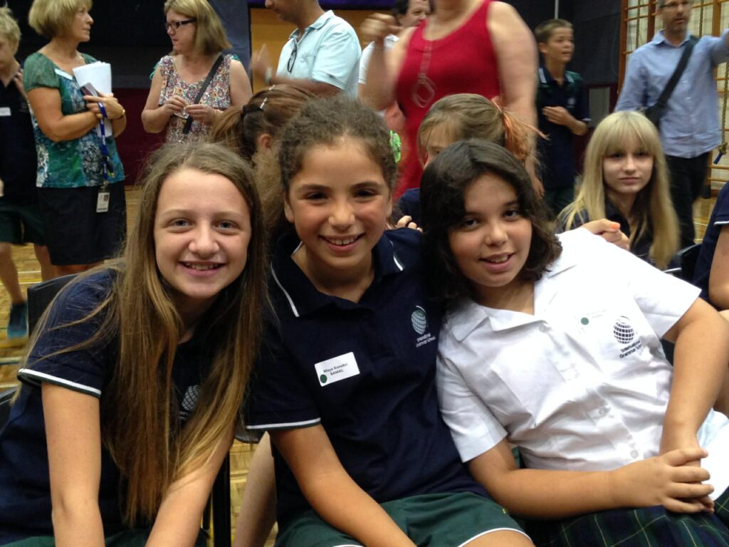 Three smiling girls in school uniforms sit together at an indoor event, with more people and students in the background—capturing a real-life Triple Play moment reminiscent of WITWAT episode 38.