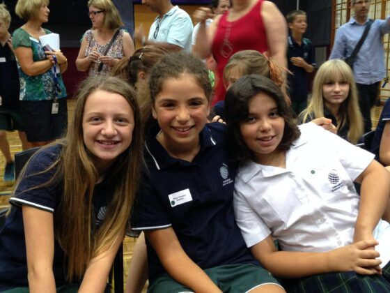 Three smiling girls in school uniforms sit together at an indoor event, with more people and students in the background—capturing a real-life Triple Play moment reminiscent of WITWAT episode 38.