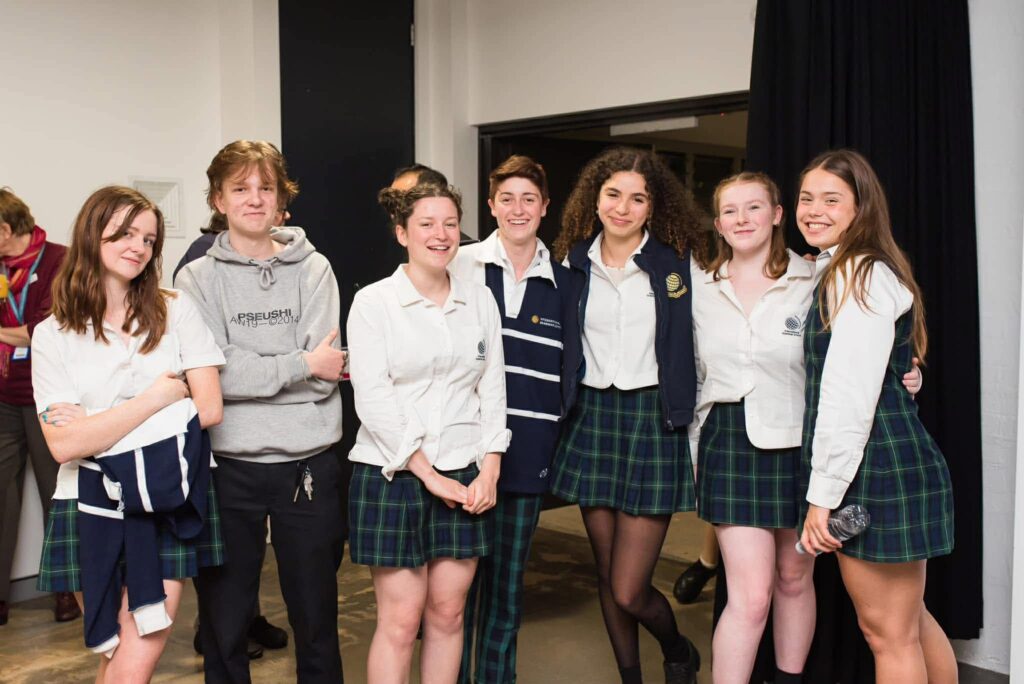 A group of seven teenagers in school uniforms stand together indoors, posing and smiling at the camera, as if ready to film episode 38 of WITWAT. Some wear white shirts; others have jumpers or jackets, and most are dressed in checked skirts or trousers.