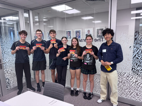 Seven students stand indoors holding colourful cards, smiling for the camera as they celebrate their achievements in Languages Beyond School. The group is casually dressed, mostly in dark clothes, and stands in front of glass walls with white patterns and framed photos.