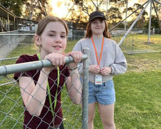 Two girls stand near a chain-link fence on a grassy field during a week of discovery. One leans on the fence, while the other, wearing a cap and lanyard, stands just behind it. Trees and metal structures are visible in the background.