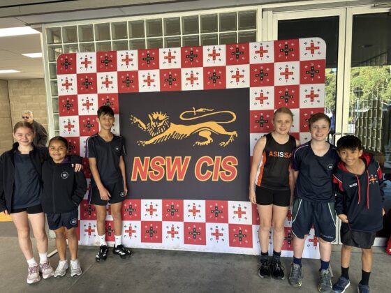 Six children in sports kits stand smiling in front of a NSW CIS sign with a gold lion logo and a red and white chequerboard background decorated with small crosses, proudly showcasing the Spirit of Athletics.