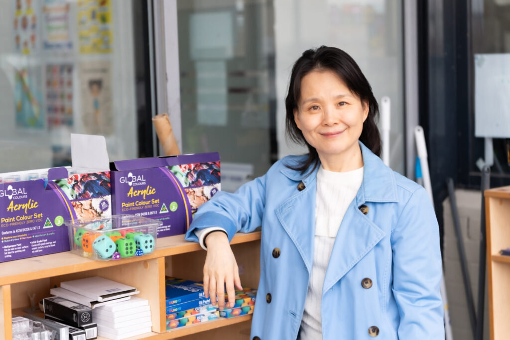 A woman in a blue coat smiles while standing indoors by shelves stocked with art supplies, colourful dice, and stationery items, embodying the spirit of Lei Feng as an Artist in Residence.