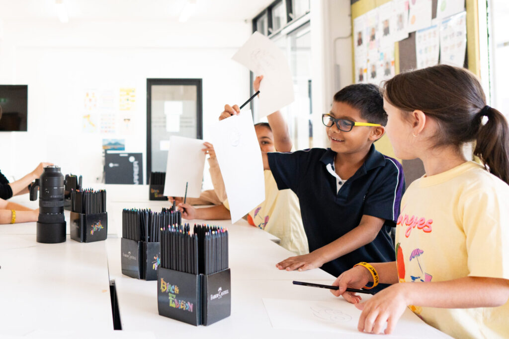 Children sit around a table in a bright classroom, holding up drawings and pencils, smiling and engaging in an art activity. Guided by an Artist in Residence, they explore their creativity together whilst sharing ideas. Art supplies and black marker boxes are on the table. Classroom walls have colourful displays.