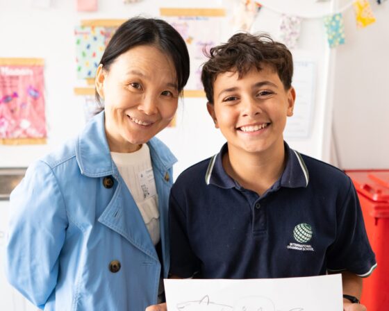 A woman in a blue coat stands smiling next to a boy in a navy school shirt who is also smiling and holding up a drawing, perhaps inspired by the school’s artist in residence, with a colourful classroom background.