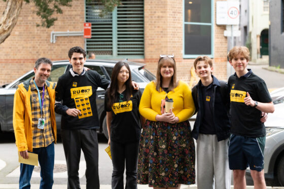 Six people stand together outdoors, smiling. Some wear black or yellow R U OK? shirts, reminding us that even a Small Question can make a big difference. A woman in the centre holds a cup and wears a floral skirt. They appear happy, standing in front of a car and brick building.