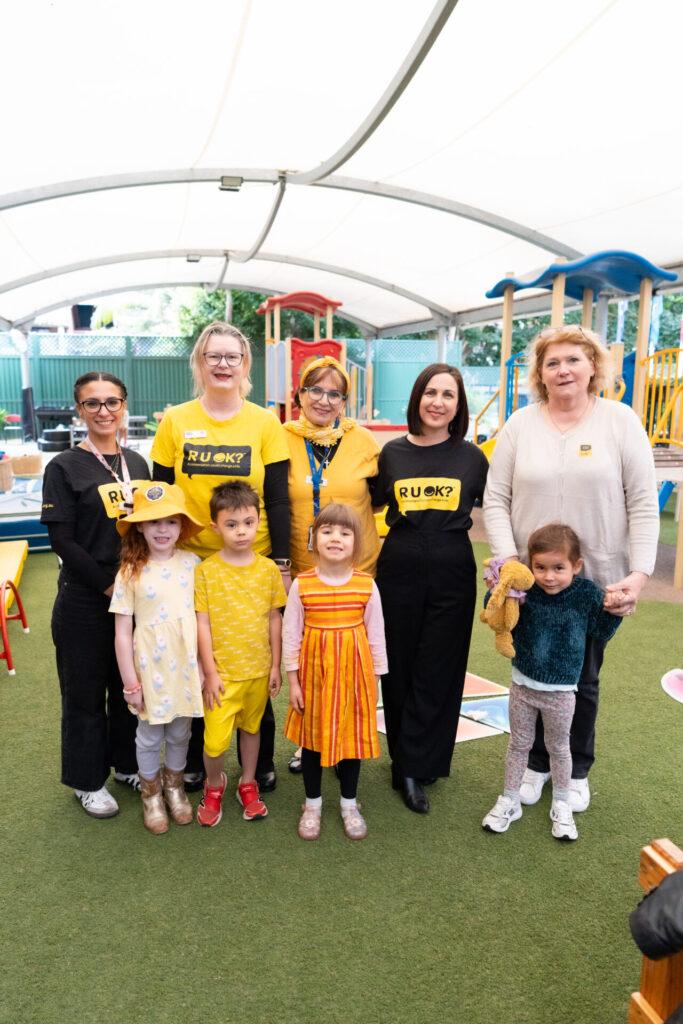 A group of adults and children, some wearing yellow clothes and R U OK? shirts, pose and smile together outdoors at a playground under a canopy, showing how a Small Question can make a Big Difference.