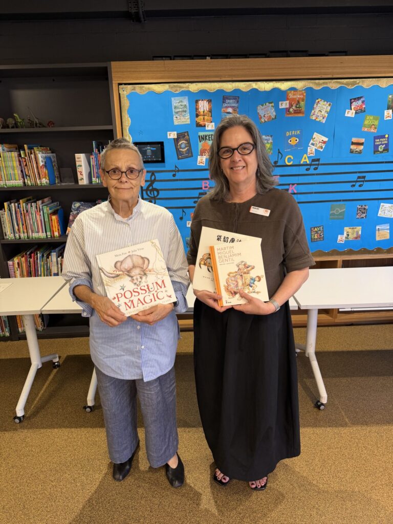 Two women stand in a library, smiling and holding children’s books. Shelves of books and a blue noticeboard decorated with musical notes are visible in the background, reflecting the spirit of learning and connection celebrated on Grand-friends’ Day.