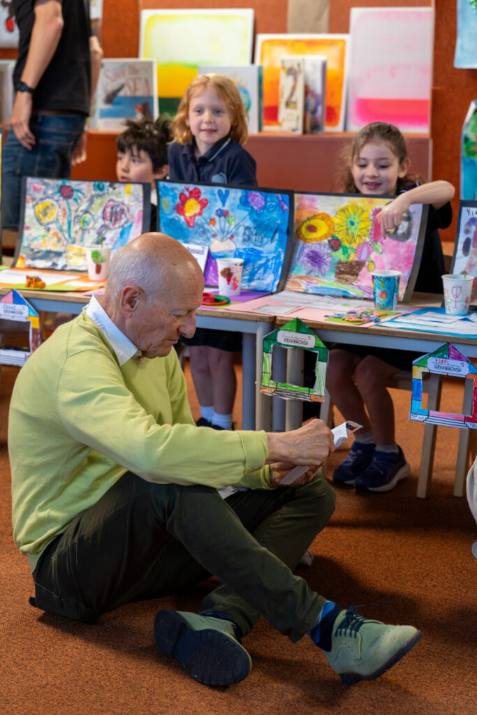 An older man sits on the floor folding paper, fostering learning and connection, whilst three young children smile behind a table displaying colourful artwork and crafts in a classroom setting.
