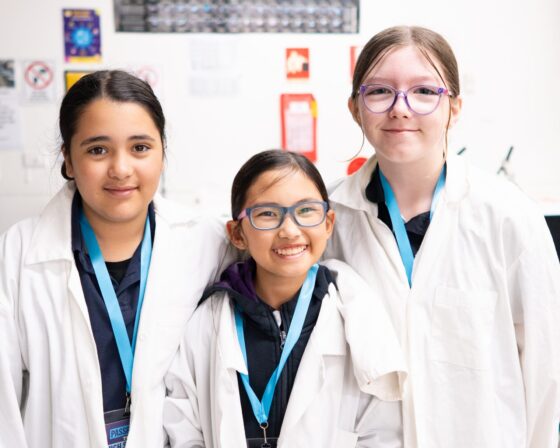 Three children wearing white lab coats and blue lanyards stand together, smiling in a brightly lit secondary school classroom or laboratory setting with science posters and red signs visible in the background, as if proudly displaying their passport to scientific discovery.
