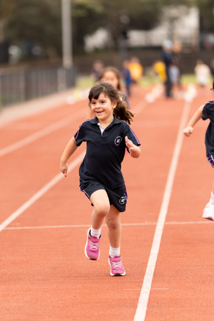 A young girl in a navy sports kit and pink trainers smiles while running on a track, embodying the Spirit of Athletics. Other children are visible running in the background.
