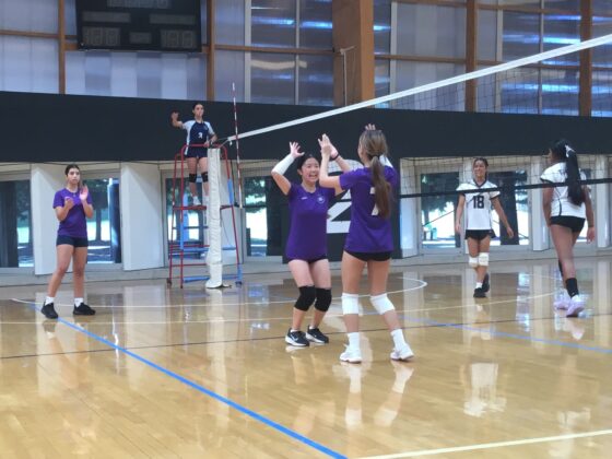 Two IGS Volleyball players in purple celebrate a point with a high five on an indoor court, whilst other players and a referee watch in the background.