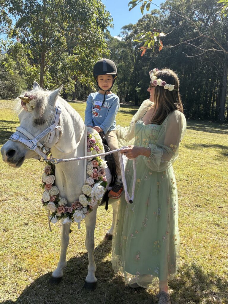 A child in a helmet sits on a white pony adorned with flower garlands, while a woman in a green dress and flower crown holds the reins during Family Fun Day at The Retreat, creating a sunny, grassy outdoor scene filled with delightful family activities.