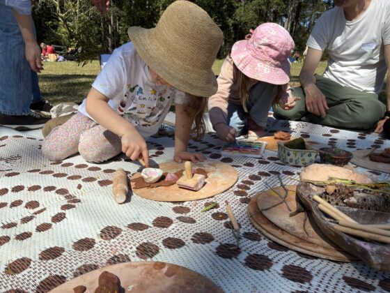 Two young children in sun hats sit on a patterned mat outdoors, using tools and clay to create art at The Retreat’s Family Fun Day. Adults and more children are nearby, with natural materials and clay objects scattered around them on the mat, making it a delightful family event for all ages.