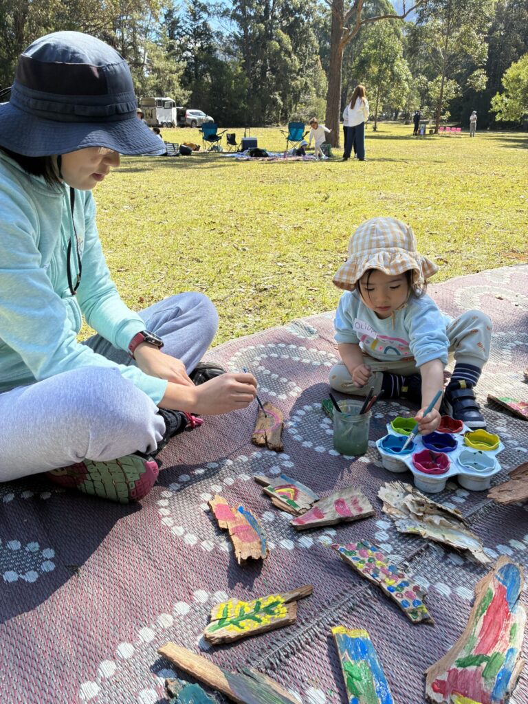 An adult and child sit on a blanket outdoors at The Retreat, painting pieces of bark with colourful paints. Trees and people are in the background on this sunny Family Fun Day.