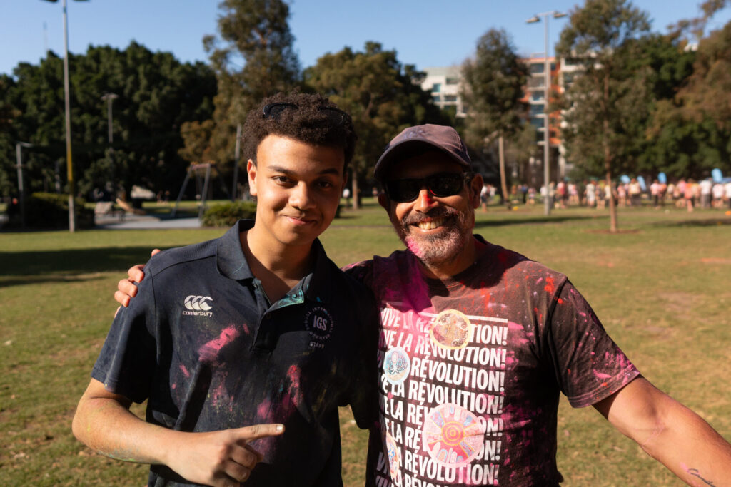 Two people smile and pose together in a sunny park, both with colourful powder on their clothes. One wears sunglasses and a hat, while the other sports a dark polo shirt. Trees and people are visible in the background.