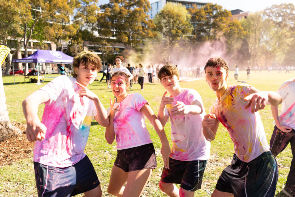 Four young people covered in colourful powder pose and smile outdoors in a sunlit park during a colour run or festival, with others and trees visible in the background.