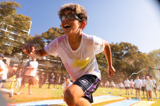 A smiling child in glasses and a white shirt runs on a colourful slip-and-slide at an outdoor event, surrounded by other children and adults on a sunny day.