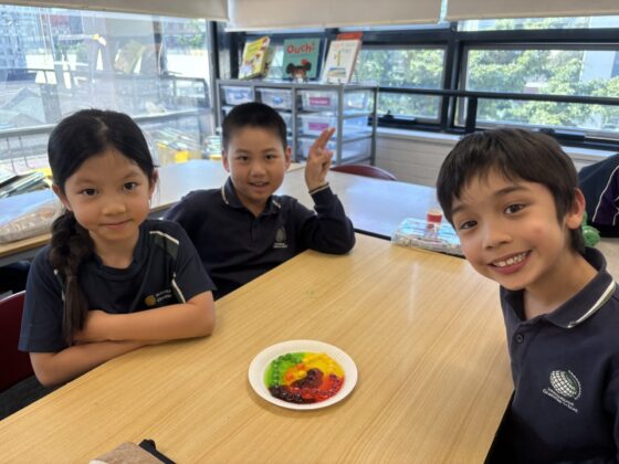 Three Year 2 children in school uniforms sit at a table, smiling at the camera. A colourful plate of sweets sits on the table as they discuss their recent language and science activities. Books and windows revealing trees and buildings are visible in the background.