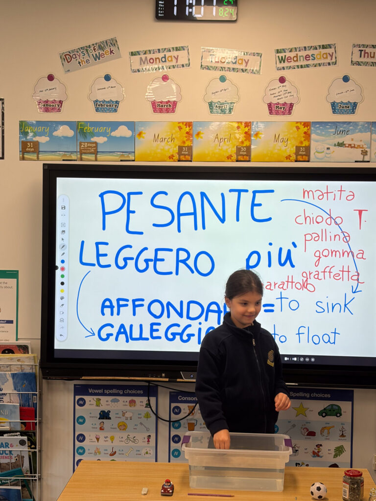 A young Year 2 pupil stands in front of a smartboard displaying Italian words and their English translations. The classroom decorations include a calendar, colourful posters, and small objects for a Language and Science lesson are placed in a clear container on the table, ready to help pupils explore water.