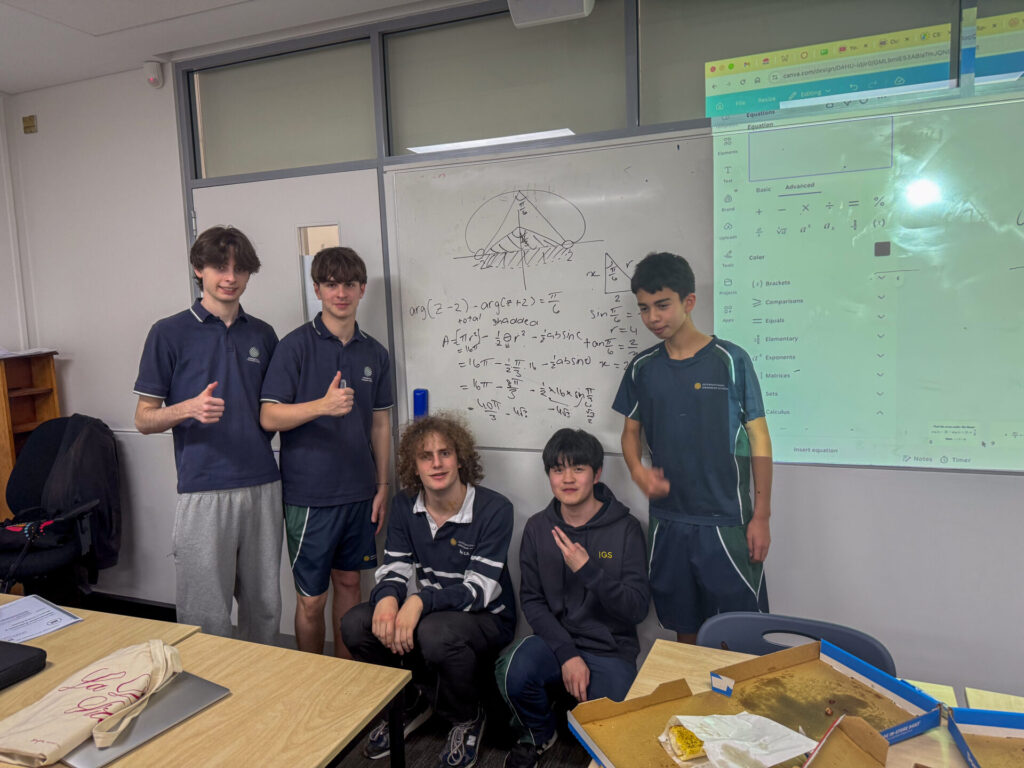 Five students pose and smile in a classroom in front of a whiteboard filled with maths equations and a drawing, commemorating their final lesson of Mathematics Extension 2. There are desks, some notebooks, food containers, and a projected screen displaying a website.