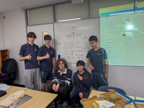 Five students pose and smile in a classroom in front of a whiteboard filled with maths equations and a drawing, commemorating their final lesson of Mathematics Extension 2. There are desks, some notebooks, food containers, and a projected screen displaying a website.