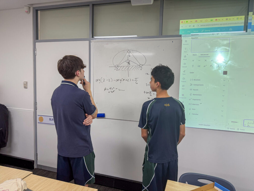 Two students in school uniforms stand at a whiteboard, discussing a challenging Mathematics Extension 2 problem involving a graph and equations. A projector displays desktop icons on the wall beside them during their final lesson.