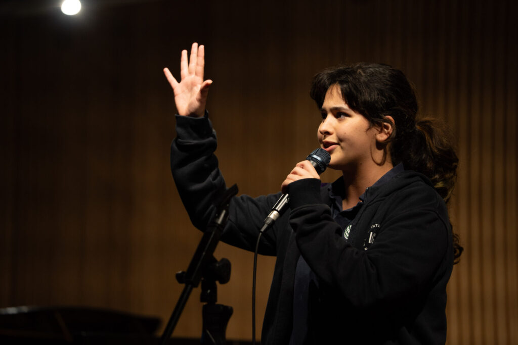 A person with dark hair holds a microphone and raises one hand whilst speaking on stage, celebrating student talent. The background is blurred and dimly lit, emphasising the speaker.