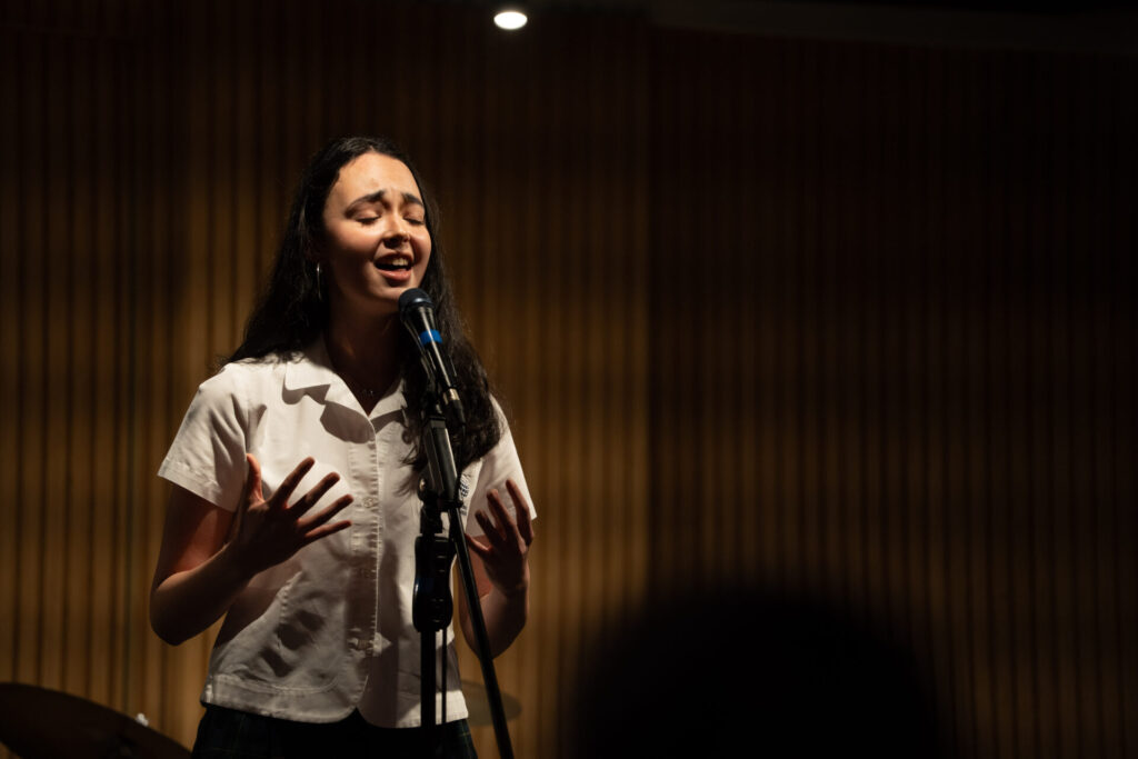 A young woman sings passionately into a microphone on stage, eyes closed and hands raised, under a spotlight with a dark, blurred background—capturing the spirit of Celebrating Student Talent.