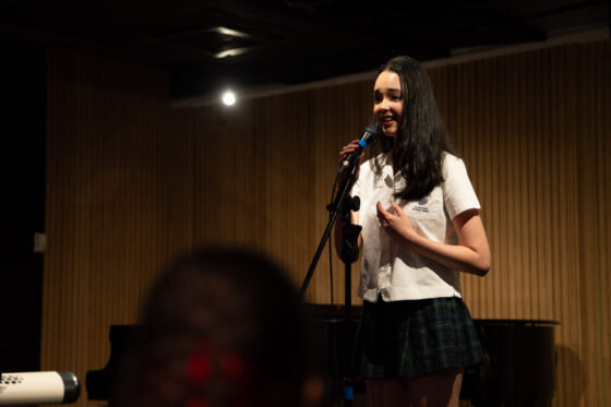 A young woman in a school uniform stands on stage, smiling and speaking into a microphone in a dimly lit room with wooden panelling and a piano in the background, celebrating pupil talent with confidence and poise.